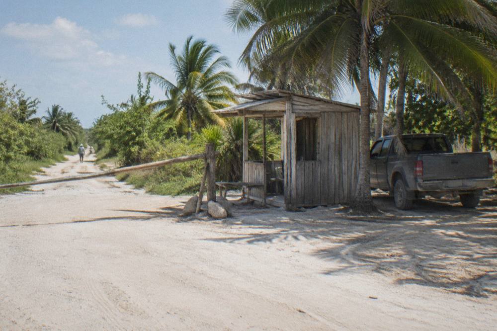 A wooden checkpoint booth beside a sandy track with palm trees and a parked pickup truck.