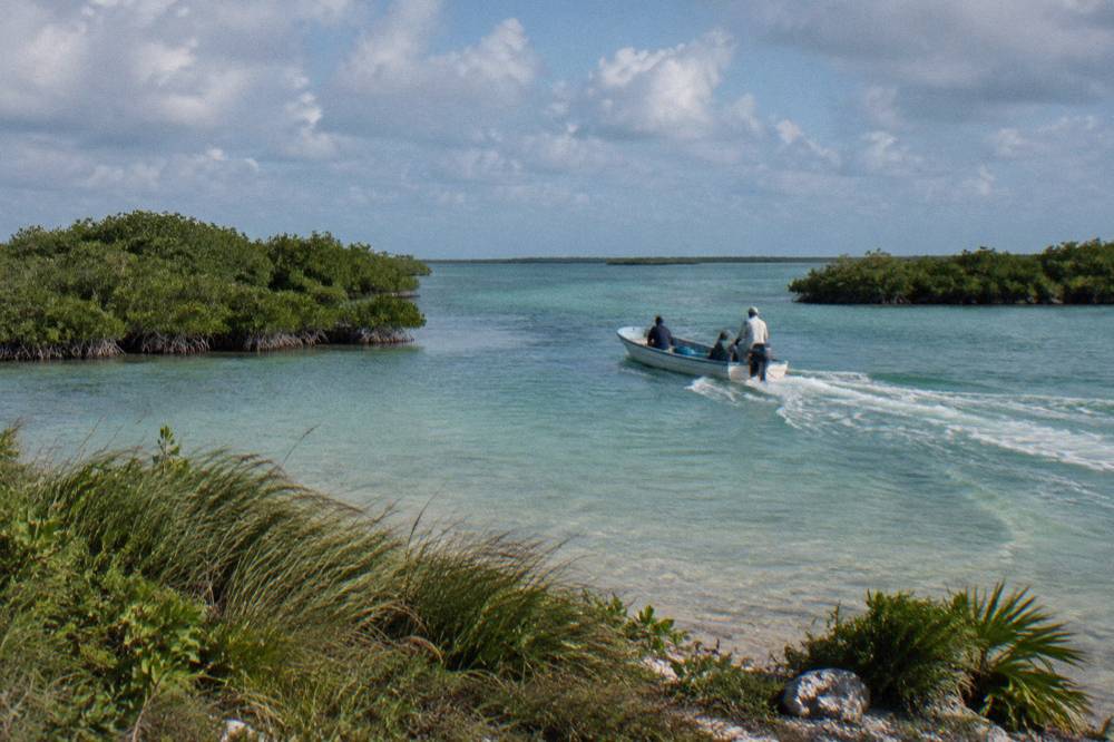 A shallow draft boat crossing a wide calm lagoon with mangrove islands under scattered clouds.