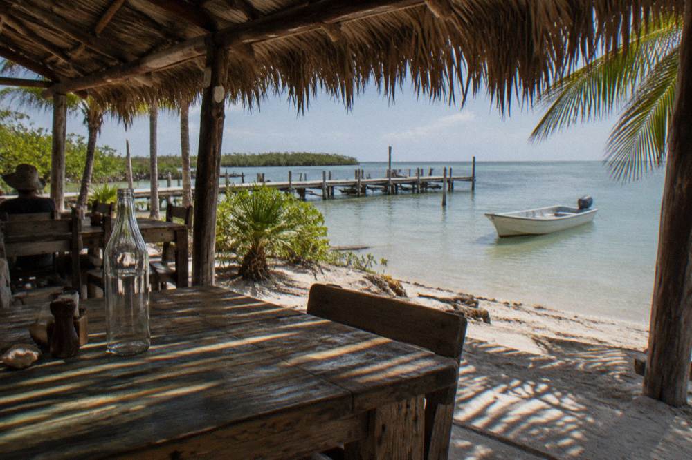 A palapa restaurant on a sandy beach with wooden tables and a moored skiff in shallow water.