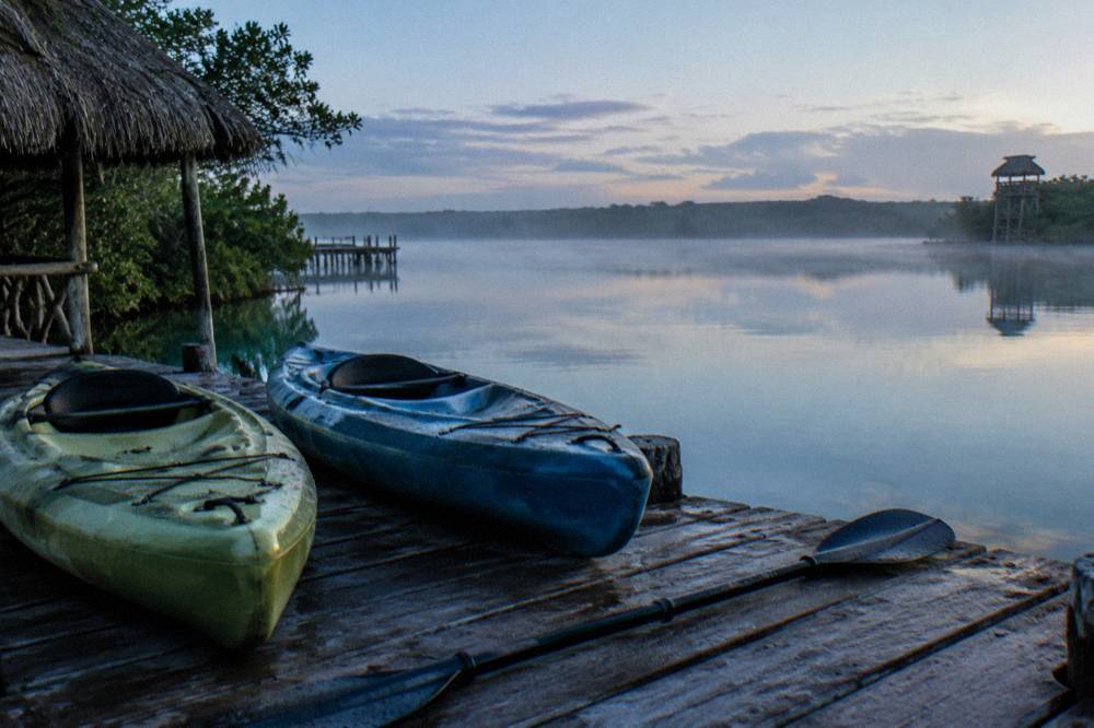 Two kayaks resting on a wooden dock beside still lagoon water at dawn.
