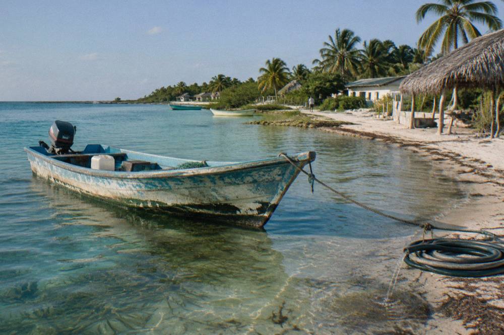 A wooden fishing boat moored in shallow turquoise water near a sandy shoreline with palms