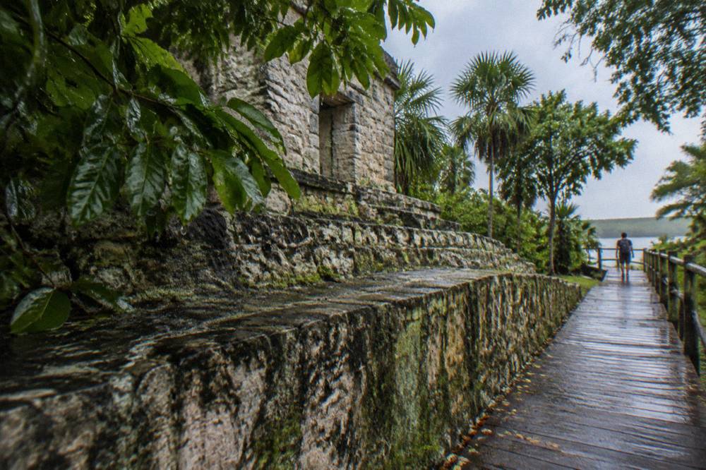 Ancient stone ruins of a small Maya temple beside a wooden walkway into green forest