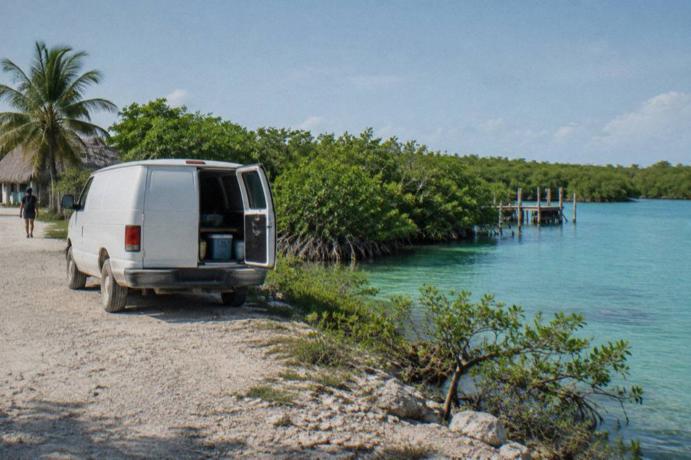 A van parked on a dirt road beside turquoise lagoon water at the edge of a mangrove forest