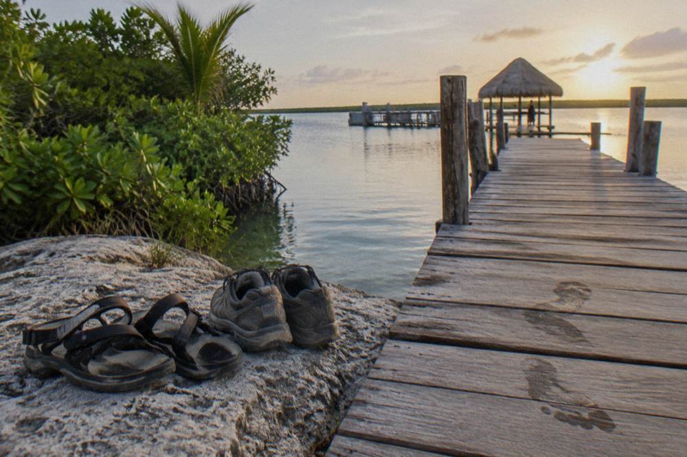 Walking shoes and strap sandals drying on a rock beside a wooden dock over still lagoon water
